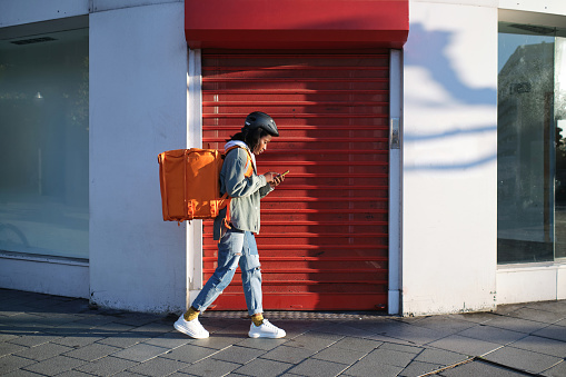 Side view of a young delivery woman with her orange backpack using her phone and walking in search of the delivery place.