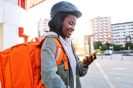 Side view of happy woman courier with thermal backpack and helmet checking order information using her smartphone.