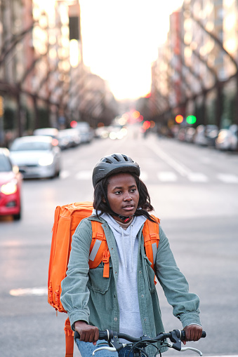 Vertical image of a young African American delivery girl riding her bike with the cars behind