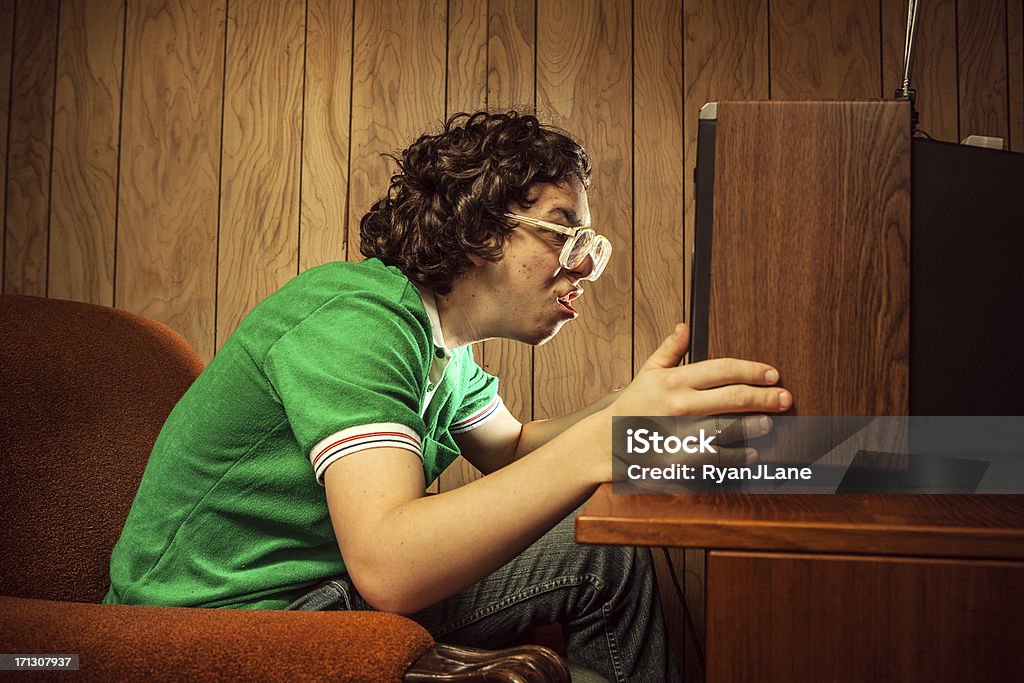 Dedicated T.V. Watcher A curly haired retro styled 1980's teenager watches something very suspenseful on the television in his living room, holding it humorously close to his face.  Horizontal with copy space on wood paneled background. Television Set Stock Photo