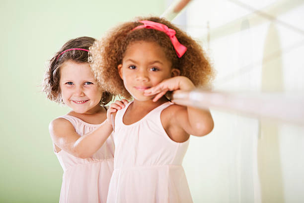 Two little ballerinas in light pink in a dance studio, holding onto a ballet barre.  The focus is on the Caucasian girl on the left; the African-American girl on the right is slightly out of focus.  Both girls wear pink headbands.