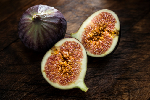 Two fresh figs, one sliced, backlit and dark against a rustic wooden backdrop.