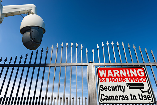 Modern security camera monitoring the entrance to a gate of a private property with a sign with text Warning 24 Hour Video Security Cameras in Use, against a clear blue sky with clouds and copy space.