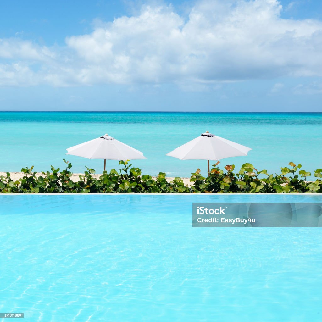 Infinity pool "Resort infinity pool in foreground with beach,  umbrellas and sea in background." Beach Stock Photo