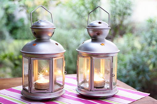 A DSLR photo of two lanterns on a table at the garden. The lanterns have burning tea lights. Front view of lanterns standing side by side. The green garden is blurred at the background. The lanterns stands on a table dressed with a pink, fuchsia pink, green and white napkin.
