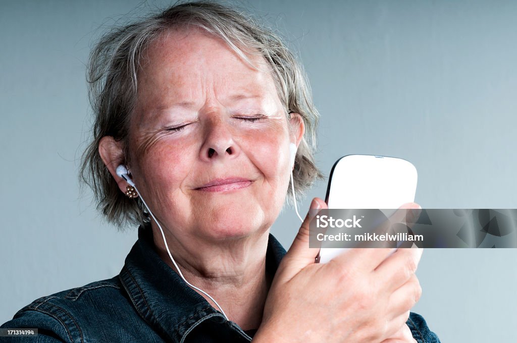 Senior woman listens to music on her phone Happy senior woman closes her eyes and listens to music, an audio book or a podcast. She wears earbuds and holds a mobile phone in her hand.  Podcasting Stock Photo