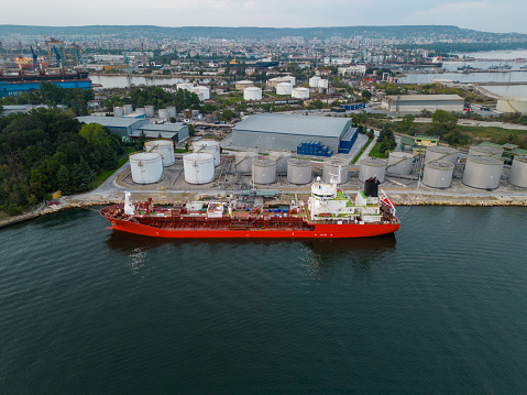 Tanker ship refueling at an oil terminal with storage silo's in the port, aerial view from flying drone