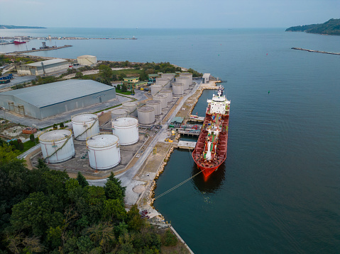 Tanker ship refueling at an oil terminal with storage silo's in the port, aerial view from flying drone