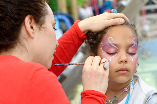 Little Girl of (6-7) is Getting Her Face Paint, Outdoors "London, UK - July 1, 2012: A little girl is getting her face painted with a butterfly. During a local summer party, kids had the chance to have a face paint session. The make up artist is carefully aplpying make up with a brush." face paint stock pictures, royalty-free photos & images