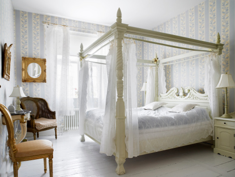 Four-poster bed in old surroundings. Pastel colors painted floorboards. Gold framed mirror on wall