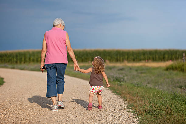 Young girl holding Grandma's hand as they walk down the gravel road at the farm. Corn field in the distance ahead of them.