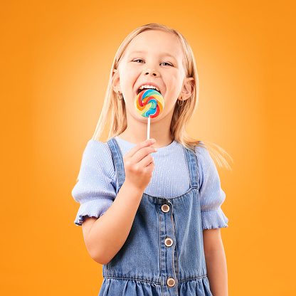 Lollipop, girl child and portrait of candy in hand, eating and studio for sweets, birthday party or carnival. Sugar, kid and lick spiral snack for dessert, hunger or meal on orange background