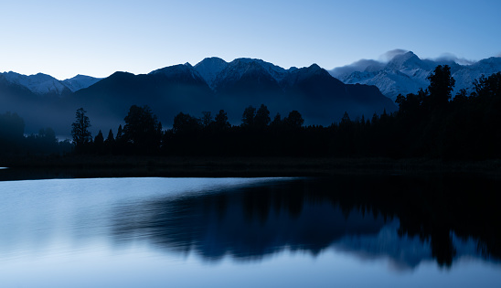 Dawn breaks over spectacular Lake Matheson in New Zealand. In the distance we see the snow capped peaks of the Southern Alps whilst in the foreground a breeze drifts across the previously mirror-like surface of the lake.