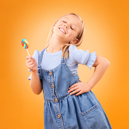 Happy, lollipop and girl child with candy, hungry for dessert  \nand isolated on an orange background in studio. Smile, sweets and young kid with sugar, unhealthy food and eating confectionery treats