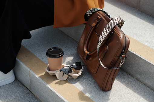 Composition of sunglasses, cup of coffee and brown leather handbag on grey concrete stairs of staircase where young female employee sitting