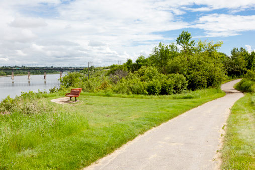 Horizontal image of paved Meewasin Trail and Bench along the South Saskatchewan River near the University of Saskatchewan. The South Saskatchewan River can be seen to the left with the CPR railway bridge visible in the distance.