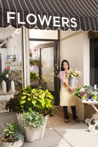 A happy Asian young woman, a Chinese Vietnamese entrepreneur business owner of an urban flower shop. The florist is standing in front of the entrance of the store holding a vase of fresh flowers in her hands, smiling, posing, looking at the camera and celebrating the opening of her business. Photographed in vertical format.