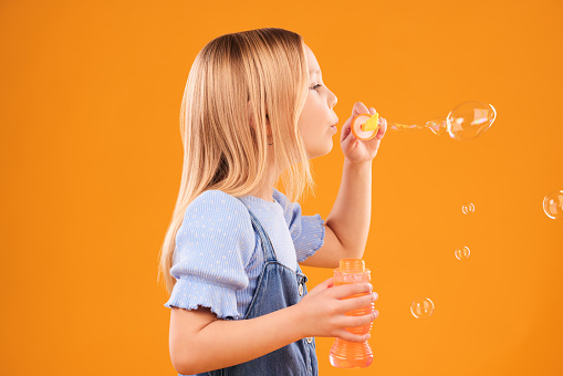 Girl, child and blowing bubbles in studio for fun, games and childhood development on mockup orange background. Profile of kid playing with soap bubble wand, toys and breathing activity for freedom