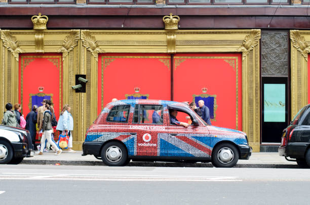 Black Cab With Vodafone Advertising "London, UK - June 2, 2012: The London's iconic black cab with Vodafone advertisement is waiting to pick up customers in front of Harrods in Brompton road.In may 2011, 100 black cabs were having this new make over. It has been designed by British designer, Christopher Kane with the collaboration of Vodafone, a mobile phone company.The design is a combination of the Union Jack flag with the prints of Kane's Spring-Summer Resort 2011 collection. Moreover, these black cabs have been fitted with mobile phone chargers and a new system that allows customers to pay their fare by text message.In the background, people are looking at Harrod's window displaying crowns designed by the world's leading brands. They were created to celebrate the Queen Elizabeth II's diamond jubilee." harrods photos stock pictures, royalty-free photos & images
