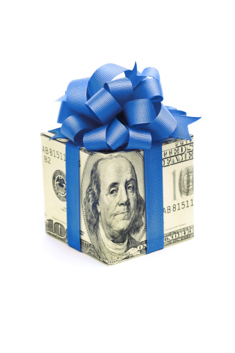 Subject: Horizontal view of a human hand holding a square gift box wrapped with paper of a U.S. one hundred dollar bill, tied with blue ribbon and a fancy bow. The bow color suggests a Hanukkah gift, bonus, birthday present, or special occasion. Isolated on a white background.