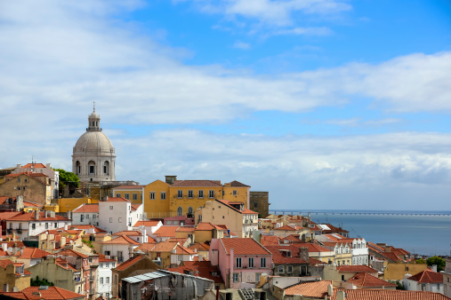 DSLR picture of the City of Lisbon in Portugal on a nice summer day. Buildings with yellow and orange roofs and walls and a church are visible. The sky is blue with few clouds.