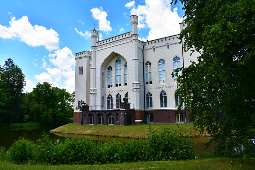 Old castle with white walls, arch and high windows. Green trees all around and blue sky. Natural historical park. Poland, Kurnik castle, Poznan, June 2022.