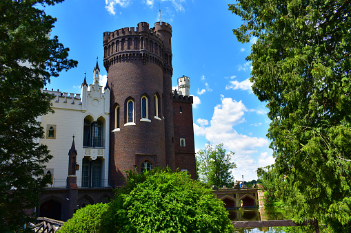 Old castle with white walls and a round tower made of dark brick. Green trees all around and blue sky. Natural historical park. Poland, Kurnik castle, Poznan, June 2022.