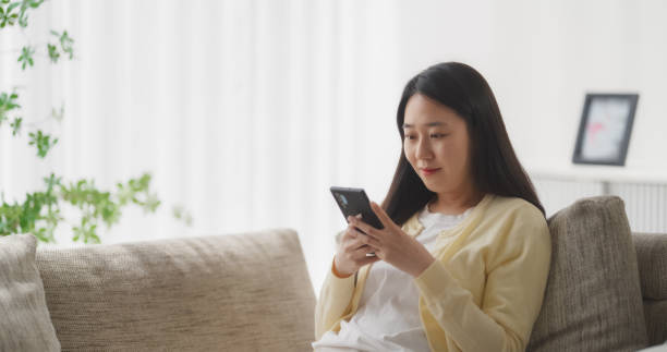 молодая южнокорейская женщина вовлечена и подключена к интернету, наслаждаясь досугом со своим смартфоном, проводя время, отдыхая на диван - проводя поиск стоковые фото и изображения