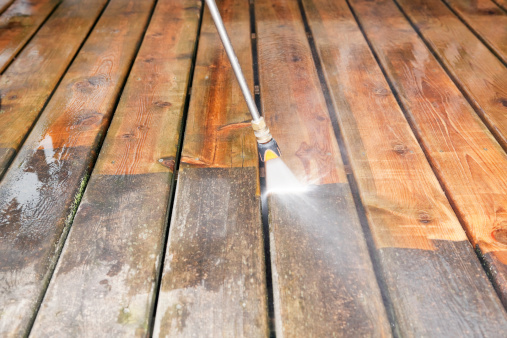 A pressure washer sprayer is cleaning a weathered treated wood deck. The background wood has been cleaned, the lower portion is dirty and weathered, showing the contrast. Focus is on the wood, the nozzle is slightly soft.