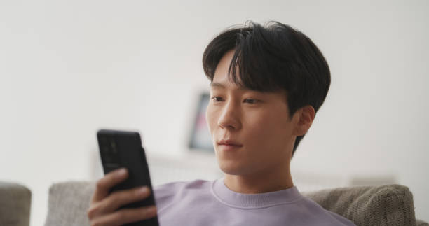 istock Young Handsome Asian Male Browsing Internet, Using Smartphone at Home, Online Shopping, Chatting with Friends on Social Media Network While Sitting on a Couch in a Living Room 1713266046