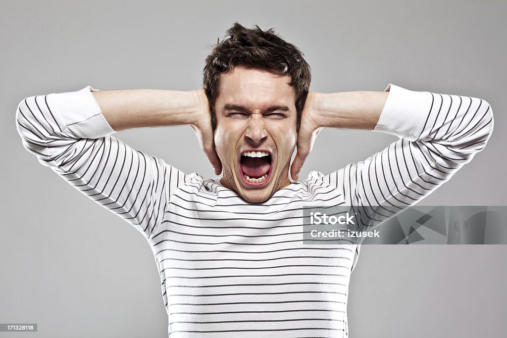 Frustration Portrait of furious young man screaming at the camera with his hands covering ears. Studio shot. Noise Stock Photo