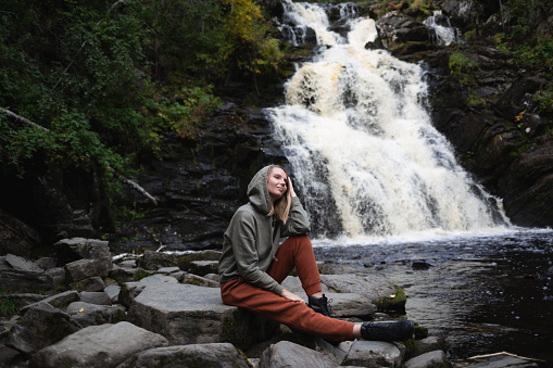 a young beautiful woman tourist sitting against the backdrop of a waterfall surrounded by forest.