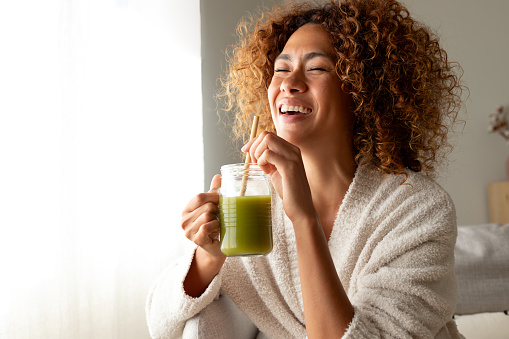 Happy multiracial woman enjoying healthy green juice at home. African American female doing detox drinking green smoothie. Copy space. Healthy lifestyle concept.
