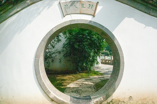 An intricately designed circular entrance in a traditional Nanjing garden, offering a peek into a serene pathway adorned with bamboo and cobblestones. An engraved wooden sign hangs above.