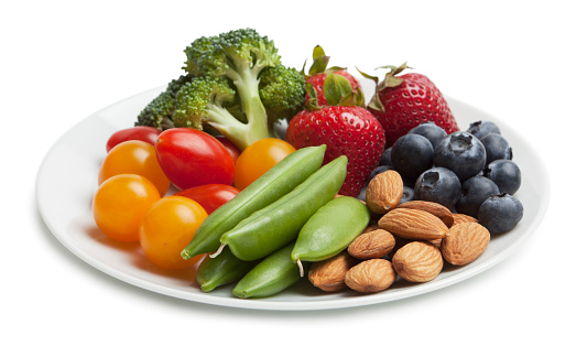 Fruit and vegetable snack plate on white background with strawberries, blueberries, almonds, sugar snap peas, tomatoes and broccoli