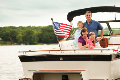 Happy, smiling American family boating on a lake motorboat, Midwest, USA. Caucasian affluent parents and children—father, mother, boy, and girl—pose for a portrait on their motorized speed boat. An American flag waves at back, celebrating a Fourth of July or Memorial Day patriotic summer vacation water sport outting.