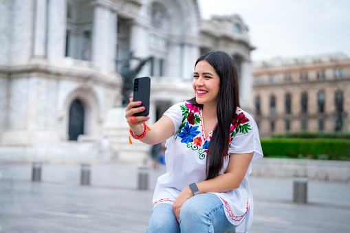 Happy young ethnic mexican woman taking selfie in front of blurred Bellas Fine Arts Palace in Mexico City