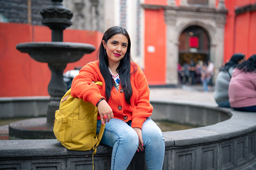Smiling young Hispanic ethnic woman sitting on fountain