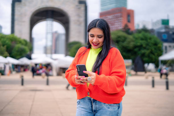 istock Smiling young Hispanic ethnic woman tourist taking using smartphone while standing in front of blurred Monument to the Revolution in Mexico City and saying hi with hand gesture in daylight 1713314372