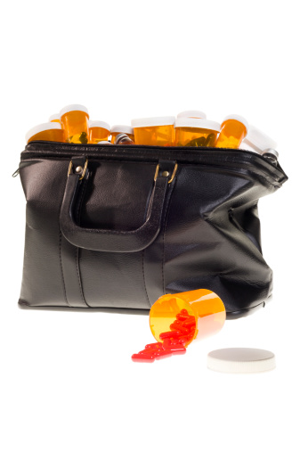 An amber pill bottle spilling red capsules in front of Doctor's Bag filled with medicine bottles on a white background