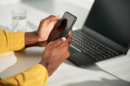 Focus on hands of black businessman with mobile phone texting or scrolling through data while sitting by workplace in front of laptop