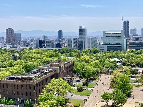 View of Osaka city from the top of Osaka Castle with green gardens, buildings, and sky, with a brown building in the foreground.