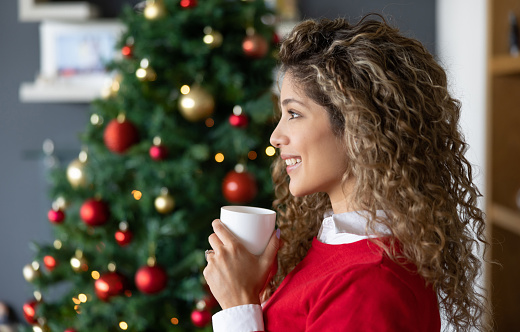 Profile portrait of a beautiful woman drinking a cup of coffee on Christmas day in front of the tree - holiday season concepts