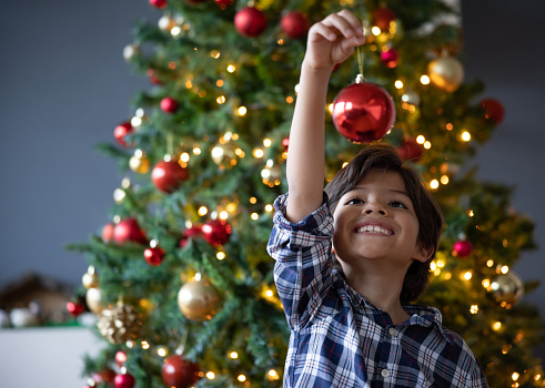 Happy boy decorating the Christmas tree and holding an ornament while smiling - holiday season concepts