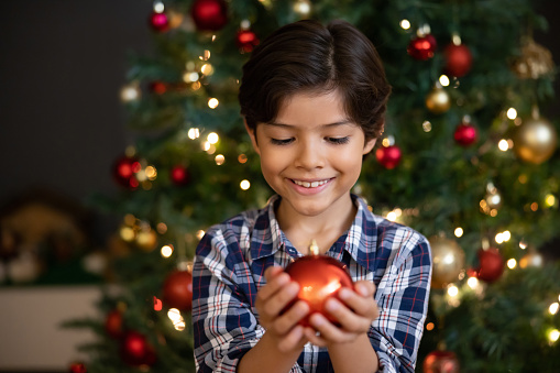 Happy Latin American boy holding an ornament while celebrating a Christmas tree - holiday season concepts
