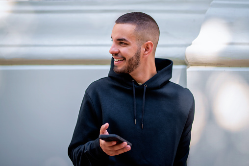 Portrait of a handsome young man using his cellphone while standing on the street in the city. Confident male wearing hoodie and text messaging.