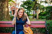 A visually impaired young woman sitting in the bench on the street and holding white cane and smartphone in her hand