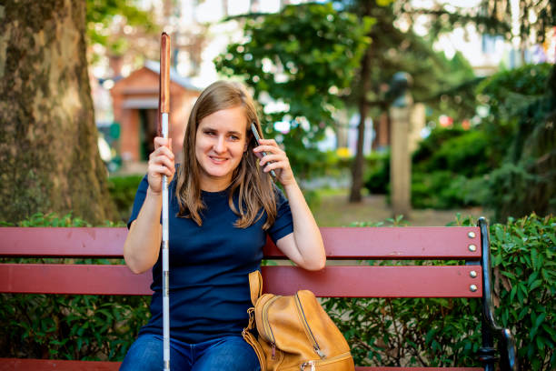 A visually impaired young woman sitting in the bench on the street and holding white cane and smartphone in her hand stock photo
