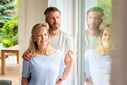 A middle-aged couple standing at the window at home. A blonde-haired woman smiling and her husband standing next to her. They wearing casual clothes.