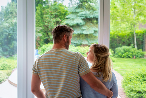 Rear view shot of couple standing together at the window and looking each other. Positive vibes.